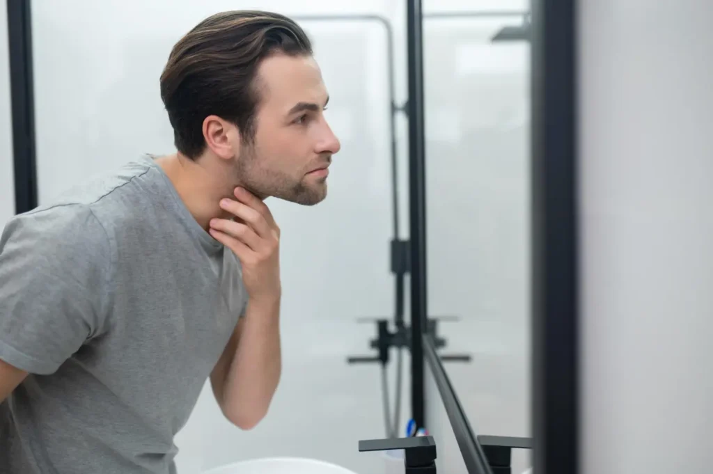 Man examining his neck and jawline in a bathroom mirror during liposuction recovery