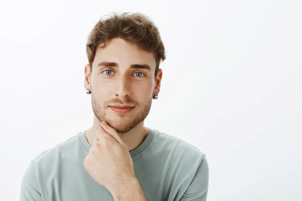 A thoughtful young man with clear skin, light brown hair, and blue eyes leans his chin on his hand