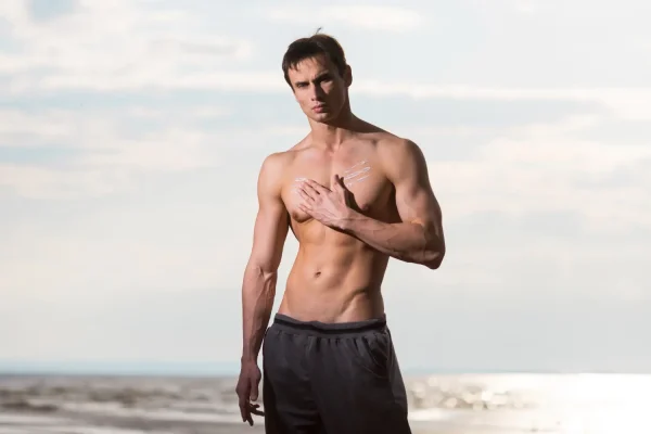 A fit man stands on a beach applying topical cream to his chest, representing a patient focused on post-operative care