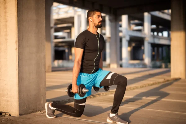 A fit man wearing earbuds performs weighted lunges with dumbbells in an urban outdoor setting