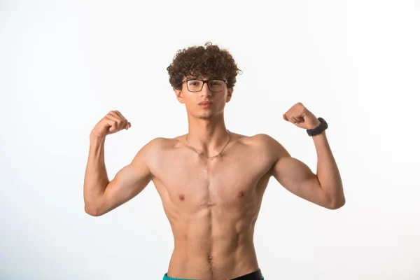 A fit young man with curly hair and glasses flexes his abdominal muscles against a white background