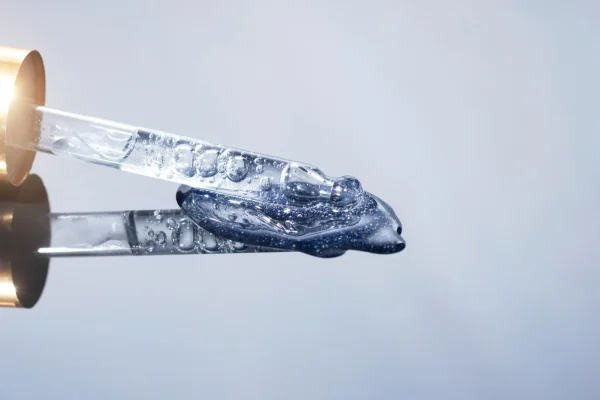 A glass dropper with a gold cap dispensing a shimmering blue-tinted serum gel