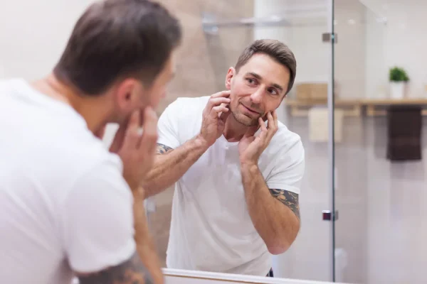 A man examines his face in the bathroom mirror, gently lifting the skin along his jawline to visualize the firming
