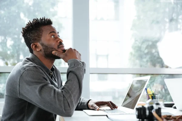 A man in a gray shirt sits at a desk with a laptop, resting his chin on his hand while gazing thoughtfully out the window