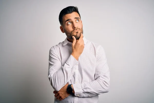 A man stands against a plain background, touching his chin and looking upward with a curious, pensive expression