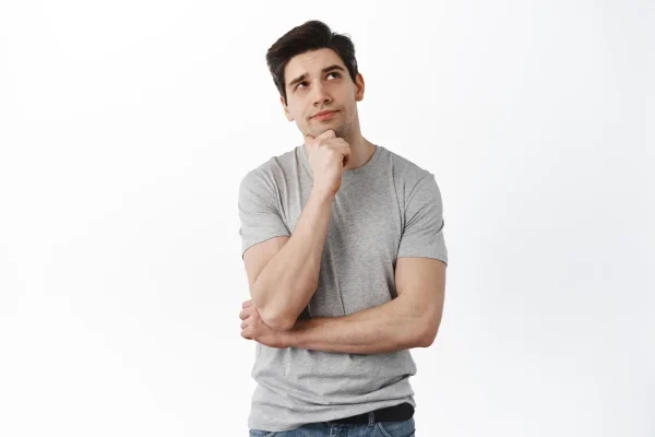 A man with dark hair in a gray t-shirt rests his hand on his chin, looking thoughtfully upwards against a white background
