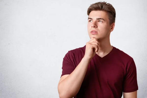 A portrait of a pensive young man in a maroon t-shirt holding his chin and looking upwards