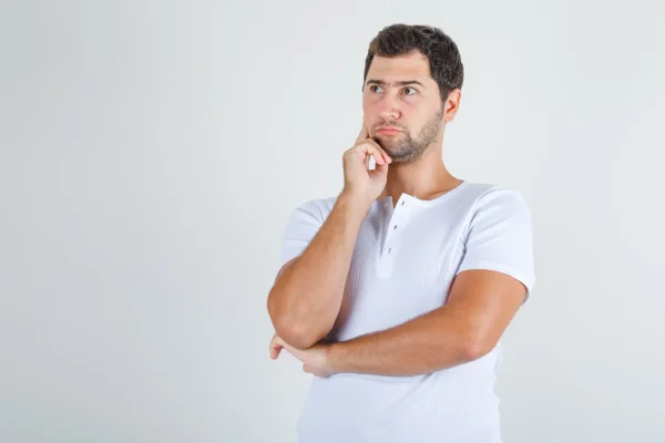 A portrait of a pensive young man with a slight beard in a white ribbed t-shirt holding his chin and looking to the side
