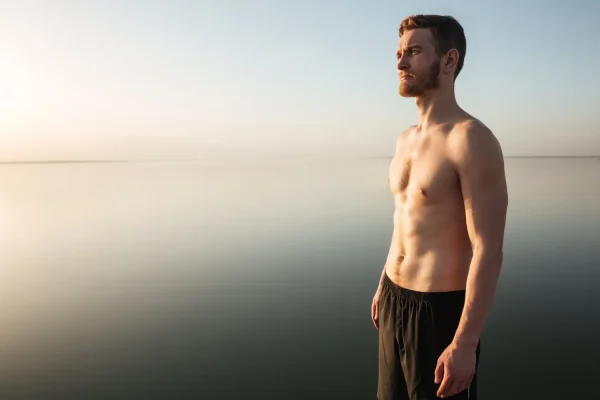 A shirtless, fit man stands by a calm lake at sunrise, looking thoughtfully into the distance