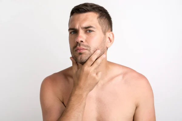 A shirtless young man carefully inspecting his jawline and neck with a concerned expression