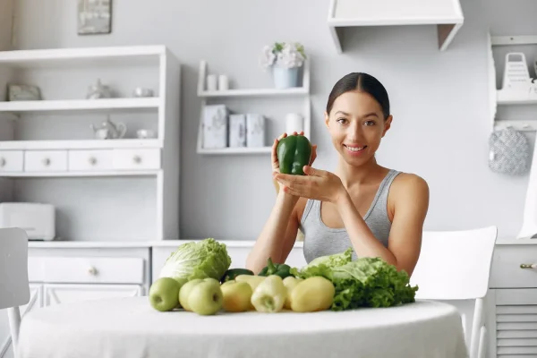 A smiling woman holds a green bell pepper while sitting at a table filled with fresh green vegetables