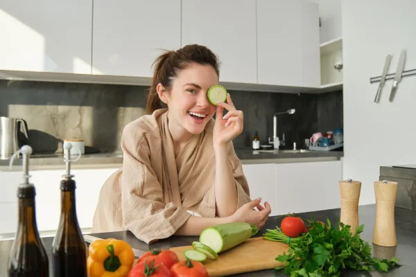 A smiling woman in a beige robe holds a cucumber slice over her eye