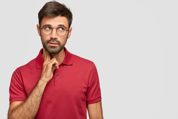 A thoughtful man with glasses and a beard in a red polo shirt looking off-camera with a pensive expression
