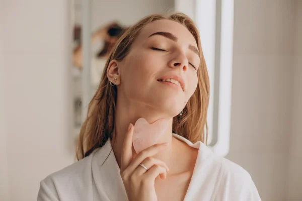 A woman in a white robe using a gua sha tool on her neck and jawline