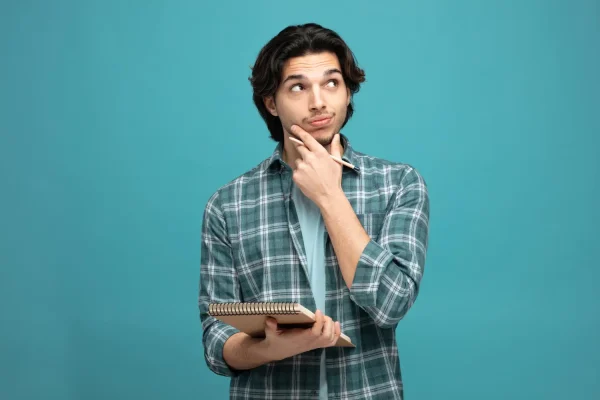 A young man in a plaid shirt holds a notebook and pencil while touching his chin in a thoughtful pose