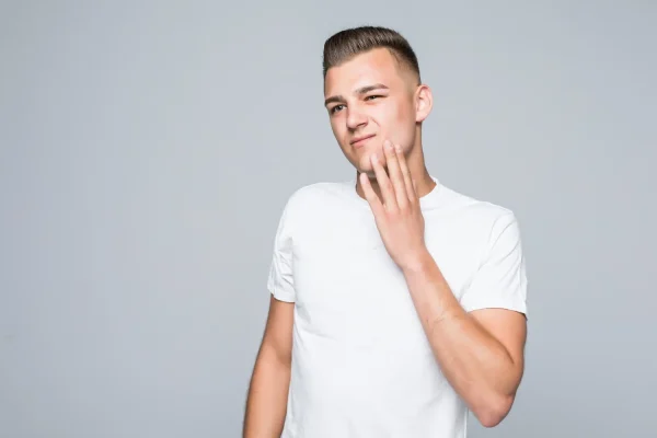 A young man in a white t-shirt gently touching his jawline with a thoughtful expression