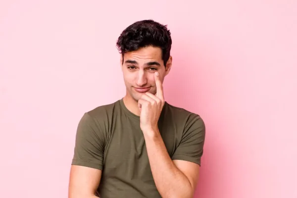 A young man points thoughtfully to the outer corner of his eye against a pink background