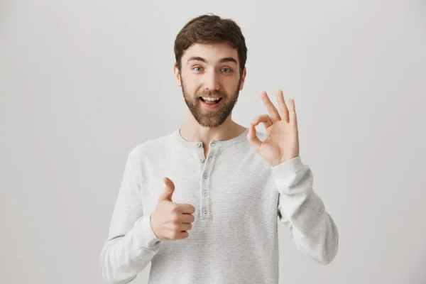 A young man with a beard smiles enthusiastically at the camera, giving a thumbs-up with one hand and an OK sign