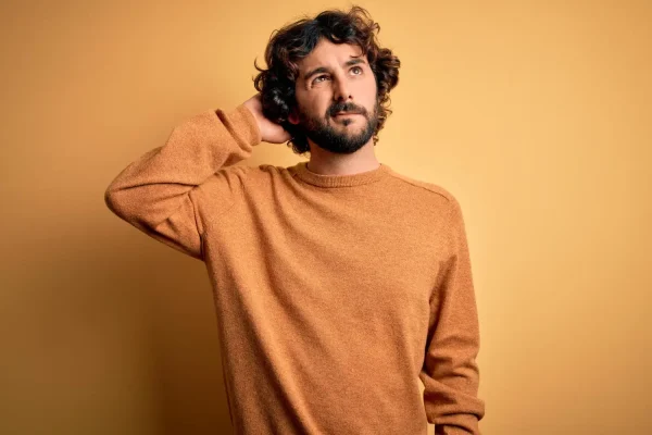 A young man with curly hair and a beard looks upward pensively while scratching the back of his head against a yellow background