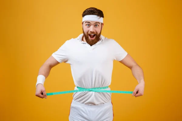 An excited man with a beard and a white headband pulls a measuring tape tight around his waist against a bright yellow background