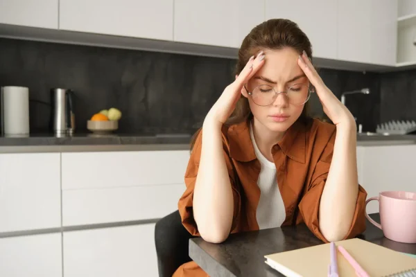 Stressed woman holding her head at a kitchen table, illustrating the connection between chronic stress