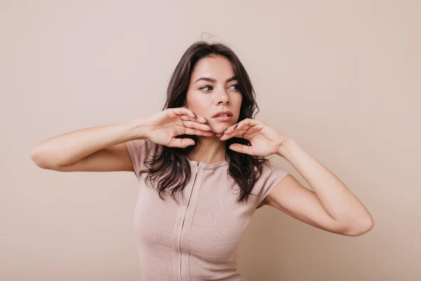 Woman with dark wavy hair pinching the skin under her chin with both hands, demonstrating stubborn neck fat that often resists diet and exercise