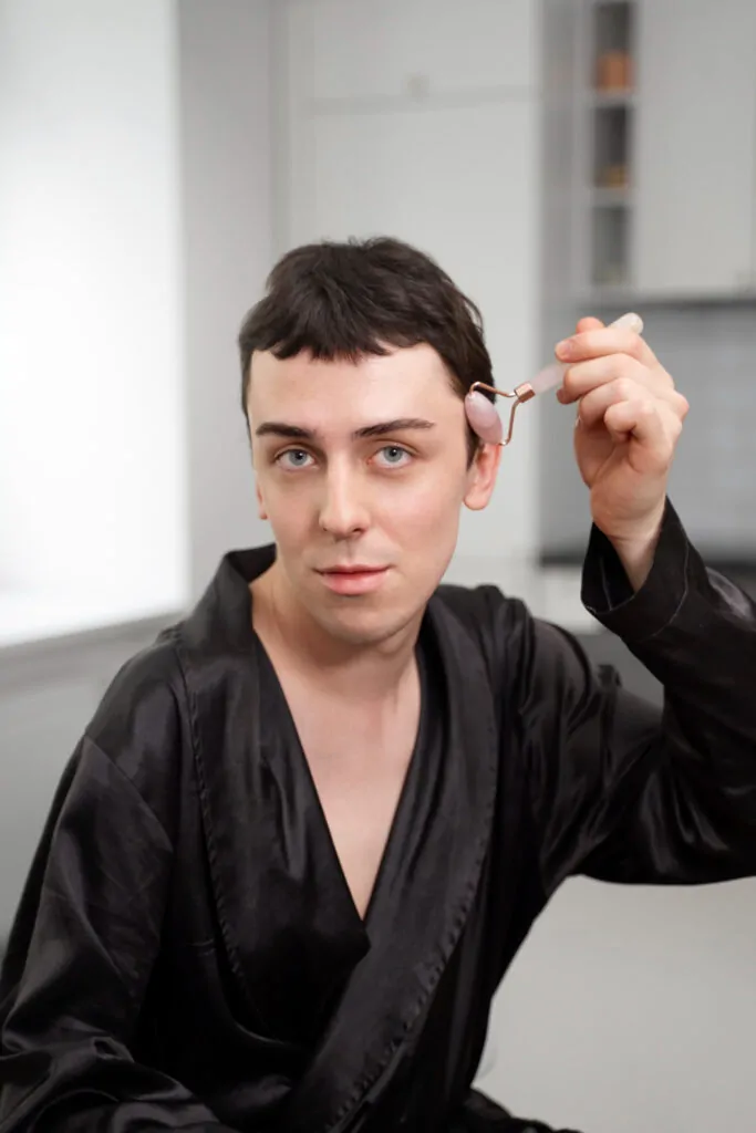 A man in a black satin robe using a rose quartz facial roller on his forehead and brow area, exploring non-surgical skincare alternatives to a plastic surgery eyebrow lift