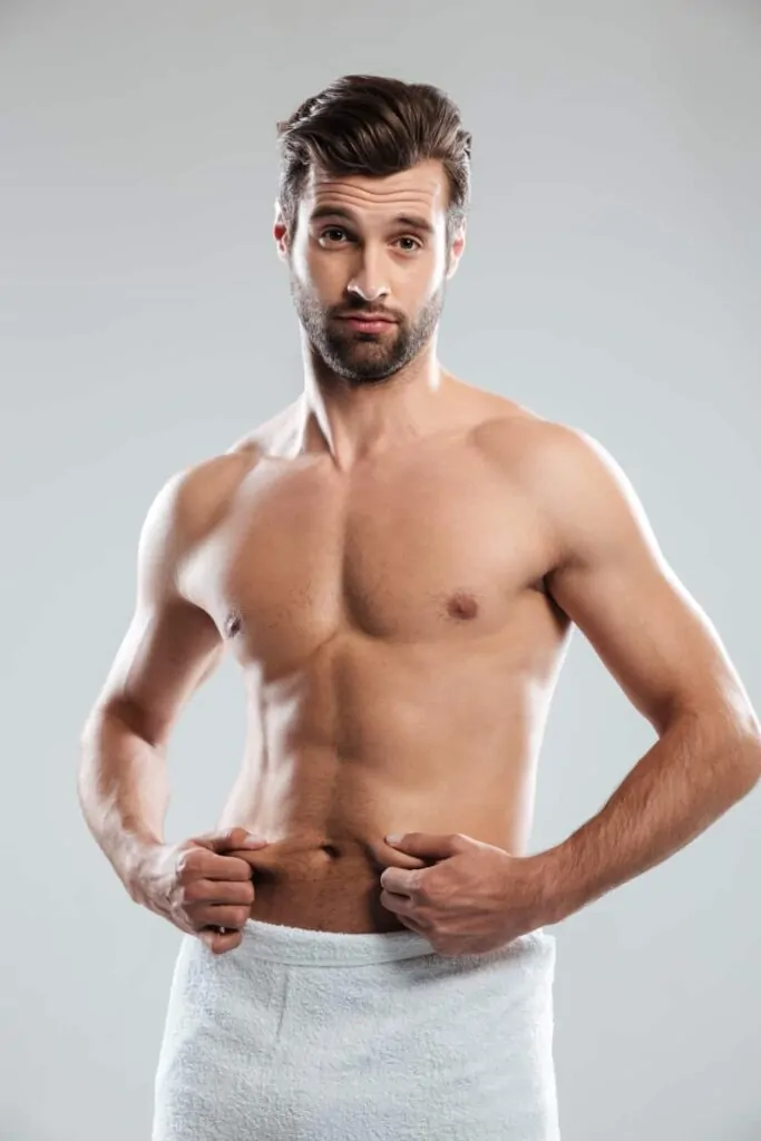 A man wrapped in a towel pinching stubborn belly fat, representing the common concern that tummy liposuction precisely and effectively addresses.