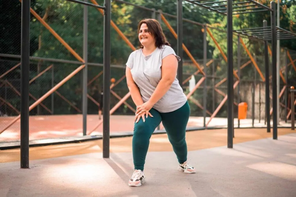 A plus-size woman in workout attire grey t-shirt, teal leggings, and sneakers smiles while stretching at an outdoor