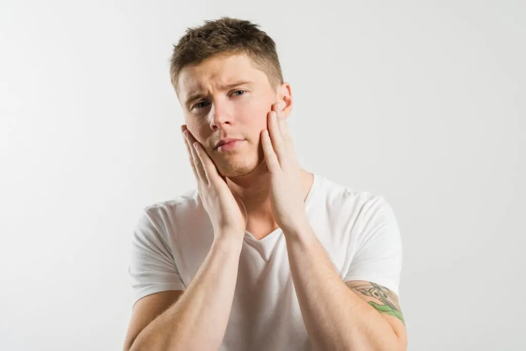 A young tattooed man in a white t-shirt cups both hands against his cheeks with a concerned expression