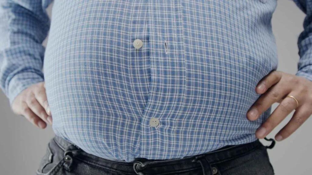 Close-up of a man's midsection showing a visibly protruding belly straining against a buttoned plaid shirt