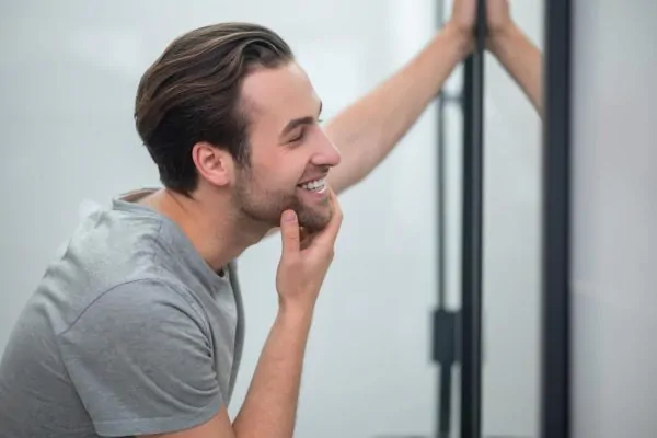 Young man in grey tshirt looking at his reflection in the mirror after getting fillers