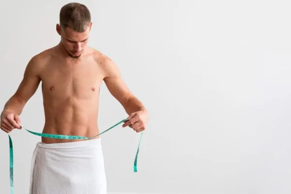 Front view of shirtless man in a towel holding measuring tape after tummy tuck