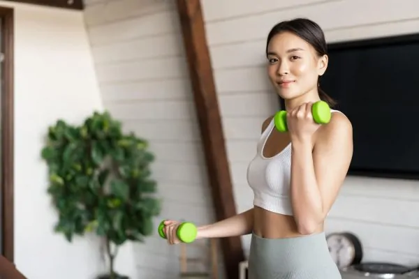 A fit woman in athletic wear exercises with light green dumbbells at home, demonstrating smart, low-impact fitness habits to maintain long-term results after neck liposuction