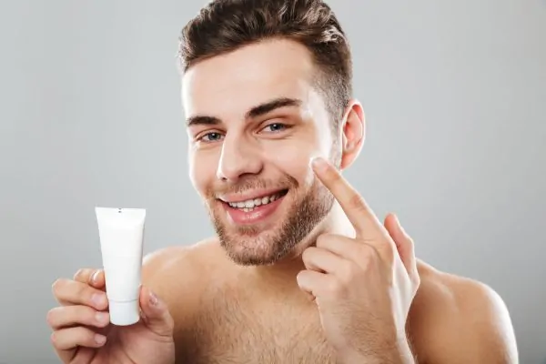 A happy young man holding a tube of numbing cream while applying it to his face, preparing for a comfortable Botox treatment