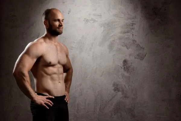 Fit man with defined chest and abdominal muscles standing against textured gray background, demonstrating body contouring results