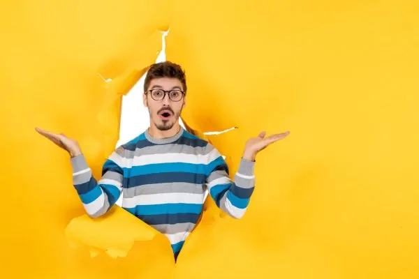 Front view stunned young man peeping through hole in paper yellow wall (1)