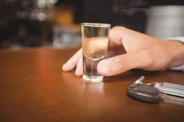 Hand holding a shot glass of clear alcohol on a wooden table with car keys nearby