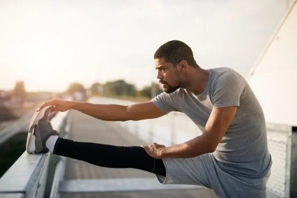 Man in athletic wear stretching his leg on a railing outdoors at sunset, demonstrating safe ways to gradually resume exercise after surgery.