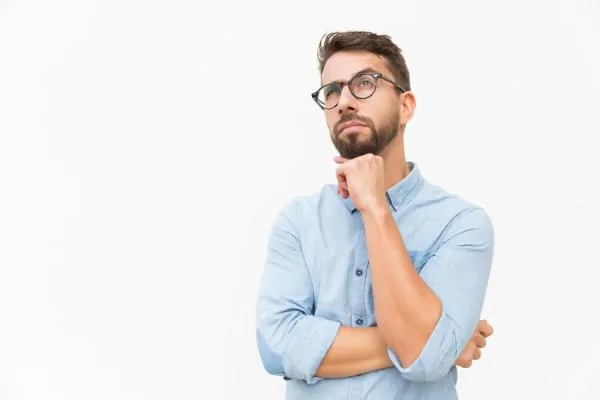 Man with glasses in a blue shirt looking upward thoughtfully with hand on chin, contemplating essential points to remember about tummy tuck surgery.