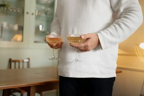 Person in beige sweater holding two champagne coupes in a home setting