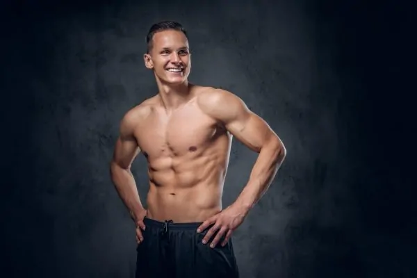 Smiling fit man showcasing well-defined six-pack abs and muscular physique against dark studio background