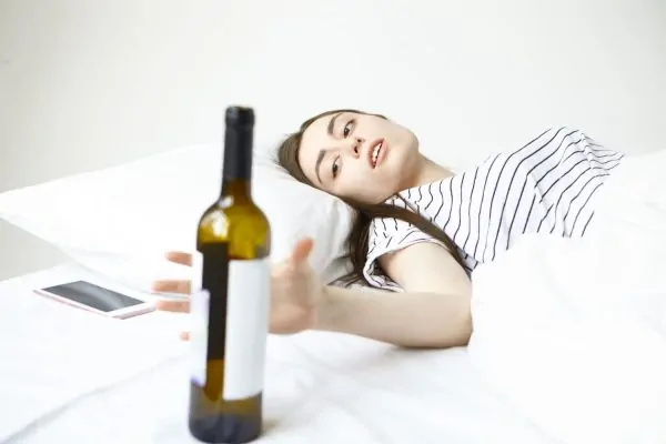 Woman lying in bed with wine bottle on table, depicting alcohol consumption that can hinder liposuction recovery