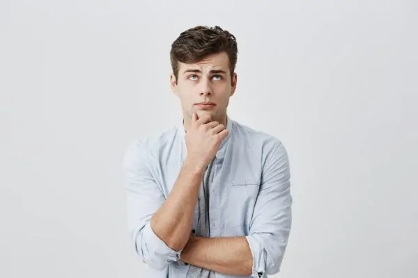 Young man with hand on chin looking upward with a curious expression, considering the differences between a full abdominoplasty and a mini tummy tuck.